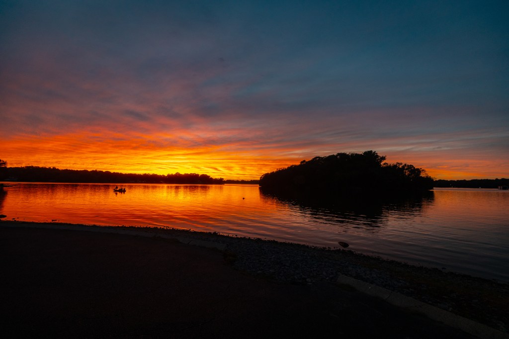 Vibrant sunset over a calm lake, with an island silhouette and clouds illuminated in orange and purple hues.