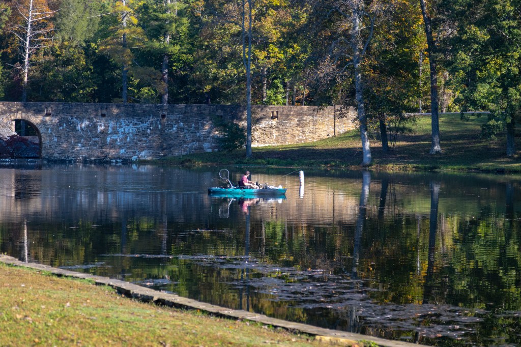 A person fishing from a small kayak on a calm lake, with trees and a stone wall in the background reflecting in the water.
