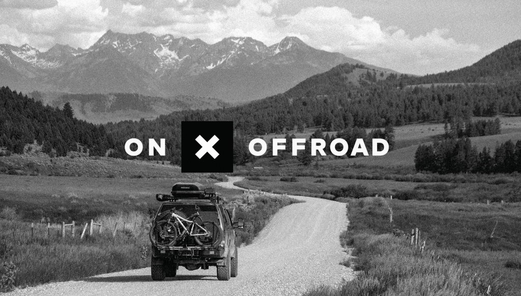 A black and white image of a dirt road winding through mountainous terrain, with a vehicle carrying bicycles on its roof near the center. The words 'ON X OFFROAD' are prominently displayed in the foreground.